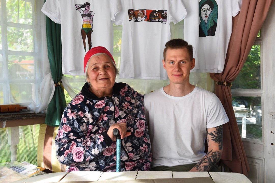 Юлия вместе с внуком Яном на фоне футболок со своими работами